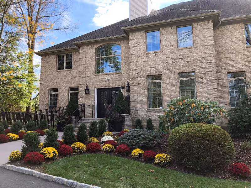 Front exterior view of a large brick building with multiple windows and a black double-door entrance. The entrance is flanked by two black lantern-style lights and surrounded by well-maintained landscaping including bushes and colorful flowers. Trees and a blue sky with some clouds are visible in the background.