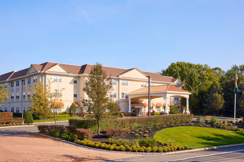 Front exterior of a three-story senior living facility with a porte-cochère entrance, landscaped grounds, and flagpoles.
