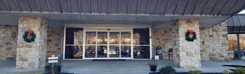 Front entrance of Deptford Center with stone pillars decorated with Christmas wreaths and glass automatic sliding doors. There are potted plants near the entrance and a small sign on the left side.