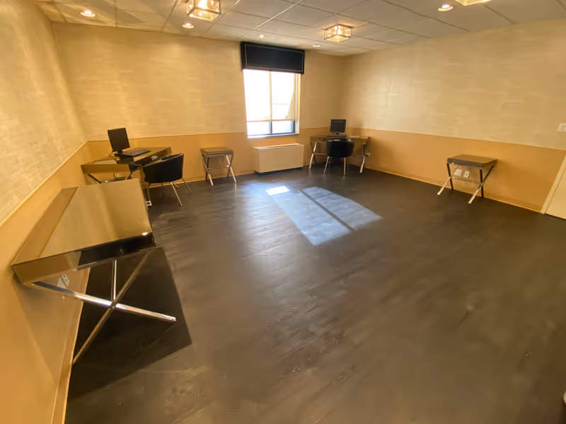 A small office or computer room with two desks, each equipped with a computer monitor and a black chair. The room has dark wood flooring, beige walls with a two-tone design, a window with a black valance, and ceiling lights.