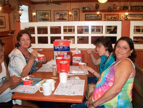 Four women sitting around a wooden table in a cozy dining area, enjoying ice cream and drinks. The table has menus, coloring sheets, and cups. The background shows framed pictures and decorative items on the walls.