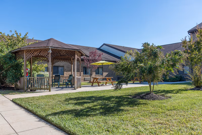 Outdoor area of a senior living facility with a wooden gazebo containing chairs, picnic tables with umbrellas, green grass, trees, and a brick building in the background under a clear blue sky.