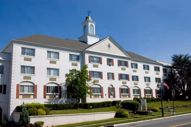 Exterior view of a large, white brick senior living facility building with multiple windows, red shutters, and a small cupola on the roof. The building is surrounded by green shrubs, trees, and a well-maintained lawn under a clear blue sky.