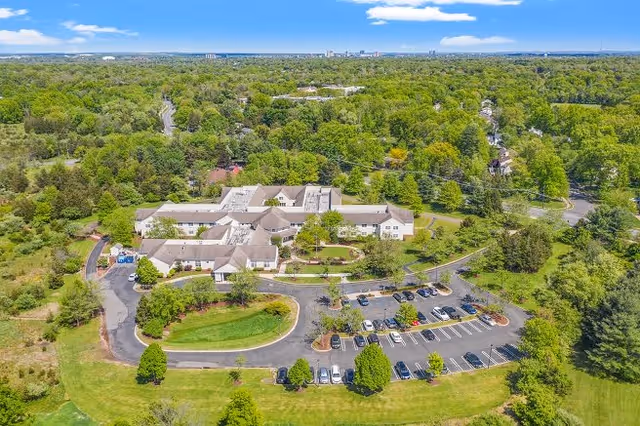 Aerial view of Spring Hills Somerset facility surrounded by lush green trees and landscaping. The building is a large, single-story structure with a circular driveway and a parking lot with several cars. The surrounding area is heavily wooded with a clear blue sky above.