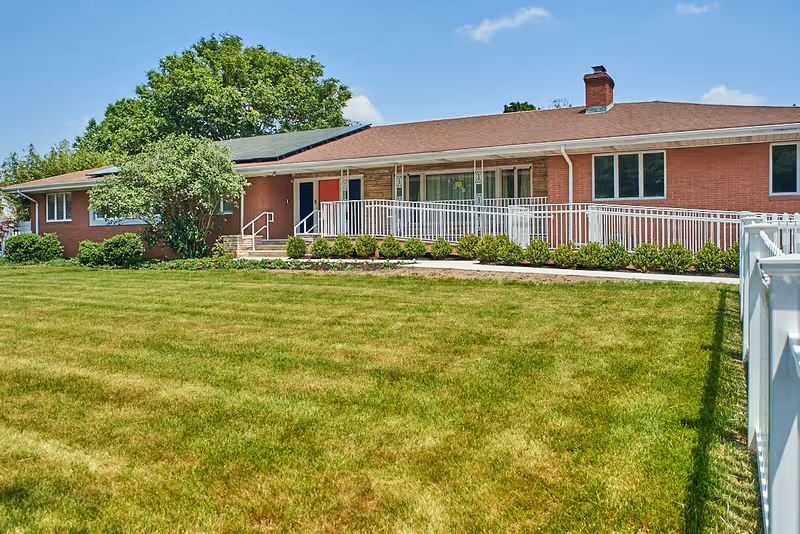 Single-story brick building with a brown roof, white railings, and a ramp leading to the entrance. The building is surrounded by a well-maintained lawn and some bushes, with a large tree in the background under a clear blue sky.