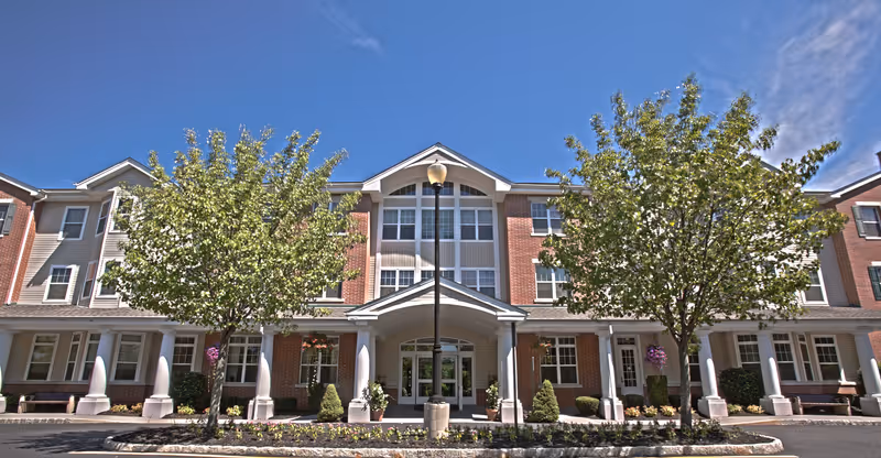 Front exterior view of a three-story senior living facility building with brick and siding facade, large windows, a covered entrance with white columns, two trees, and a streetlamp in front under a clear blue sky.