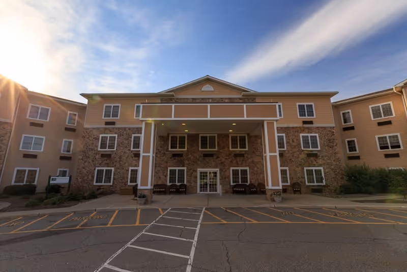 Front exterior view of a three-story senior living facility building with a covered entrance supported by two large columns. The building features a combination of stone and beige siding with multiple windows. There are chairs arranged near the entrance and a parking area with marked spaces in front. The sky is partly cloudy with the sun shining from the left side.