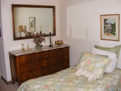 A bedroom with a wooden dresser topped with decorative items including a lamp, candles, and a plant. Above the dresser is a large mirror. Next to the dresser is a bed with floral patterned bedding and multiple pillows. A framed picture hangs on the wall above the bed.