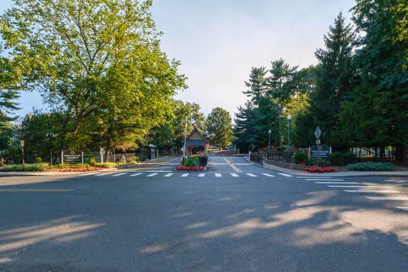 Gated entrance and guardhouse of Winchester Gardens surrounded by trees, landscaped beds, and a crosswalk.