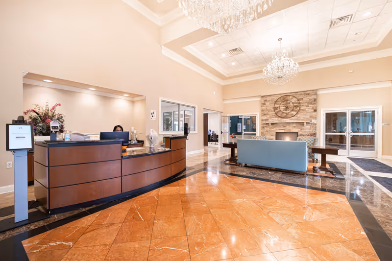 A spacious and well-lit reception area with a curved wooden front desk where a receptionist is seated. The floor is tiled with a combination of orange and black marble. In the background, there is a seating area with a blue couch facing a stone fireplace adorned with a large decorative clock. The ceiling is high with elegant chandeliers hanging down, and there are glass doors and windows providing additional light.