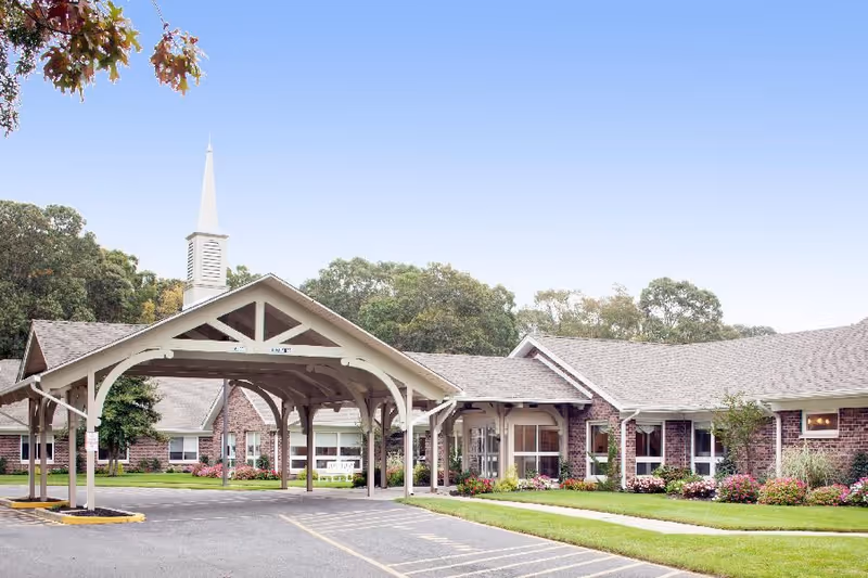 Front entrance of a single-story brick nursing facility with a covered drive-through porte-cochère, landscaped lawns, and a steeple.