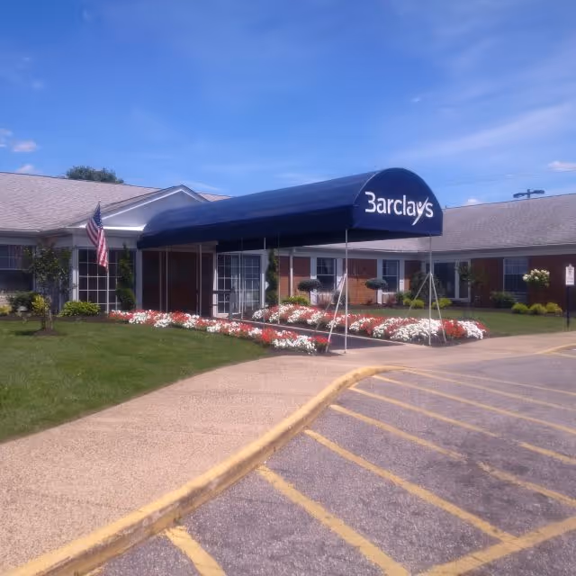 Exterior view of Barclays Rehabilitation And Health Care Center showing a single-story brick building with a blue awning entrance labeled 'Barclays'. The entrance is surrounded by well-maintained flower beds with red and white flowers, green grass, and an American flag on a pole. The sky is clear and blue.