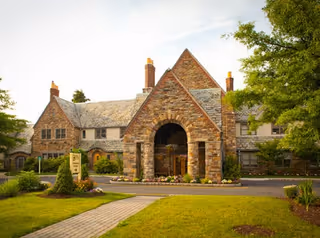 Front exterior view of a large stone building with a steep roof and multiple chimneys, surrounded by green lawns, trees, and a paved walkway leading to the entrance.