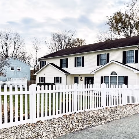 A two-story white residential building with black shutters behind a white picket fence. The building has a dark roof and several windows, with a smaller white building visible in the background. The sky is cloudy and there are some leafless trees around.