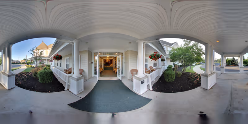 Covered entrance area of a senior living facility with white columns, hanging flower baskets, and a green entrance mat leading to double glass doors. There are landscaped garden beds with bushes and flowers on either side, and a paved driveway and walkway visible under a clear sky.