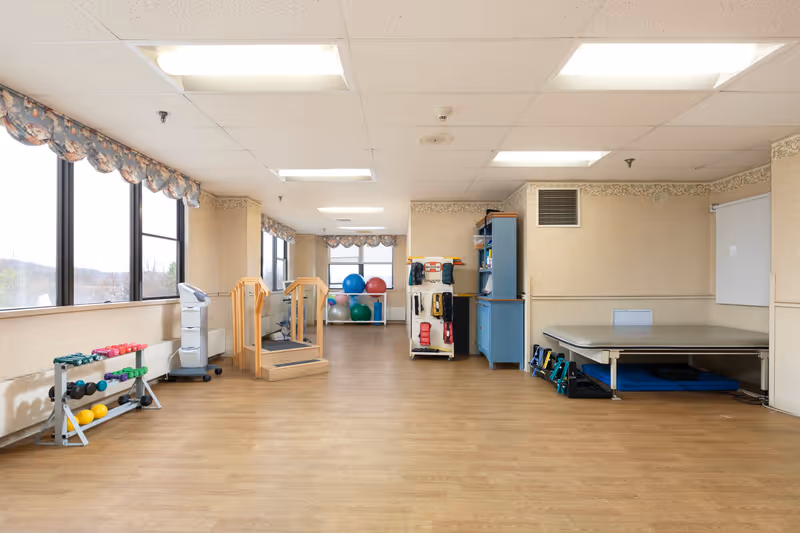A spacious physical therapy or exercise room with large windows along one wall, wooden flooring, and various exercise equipment including dumbbells, therapy balls, a wooden staircase for rehabilitation, and a padded therapy table. The room is well-lit with ceiling lights and has floral valances on the windows.