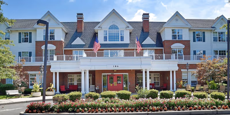 Front exterior of a large brick-and-white senior living building with a covered entrance, American flags, and landscaped flowerbeds.