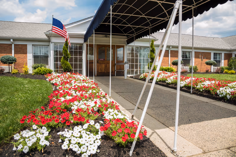 Entrance to a brick building with a covered walkway. The walkway is flanked by flower beds filled with red and white flowers. An American flag is displayed on a pole near the entrance. The sky is partly cloudy.