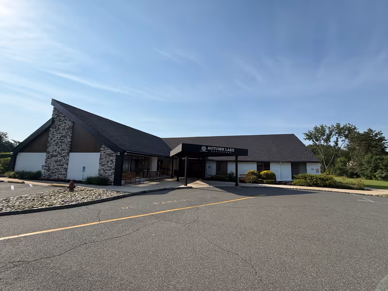 Exterior view of Autumn Lake Healthcare at Berkeley Heights building with a peaked roof, stone and white walls, a covered entrance, and surrounding greenery under a clear blue sky.