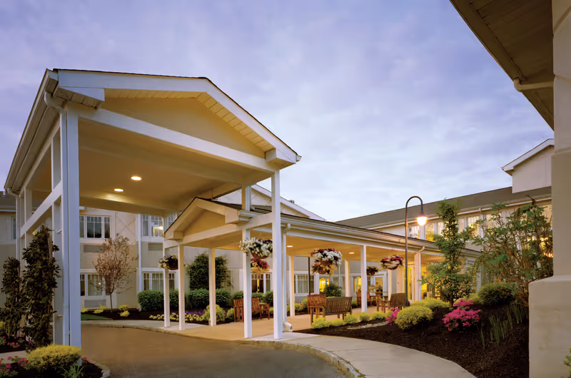 Covered entrance area of a senior living facility with white pillars and hanging flower baskets. There are benches and landscaped bushes and flowers around the walkway leading to the building. The sky is cloudy and the building has multiple windows with lights on inside.
