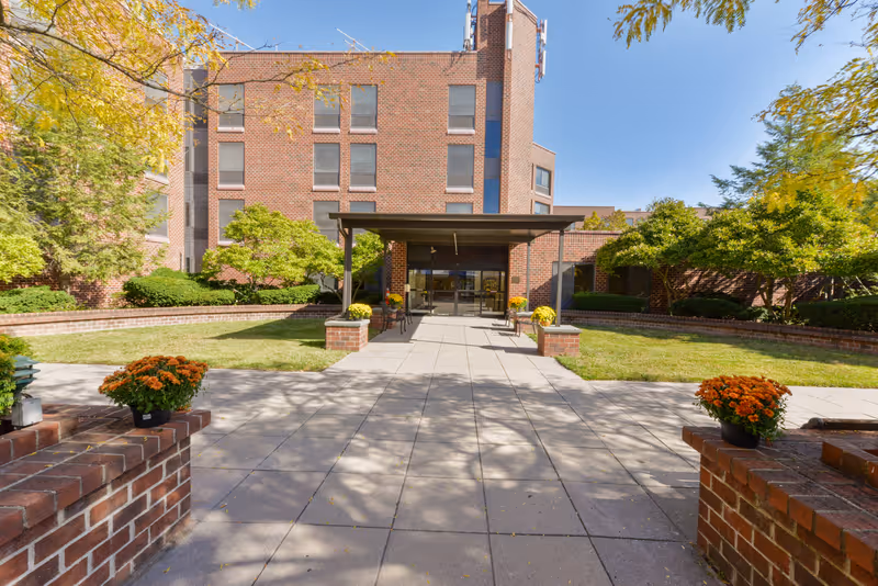 Front entrance of a brick building with a covered walkway, surrounded by green bushes, trees, and potted orange flowers on brick pillars under a clear blue sky.
