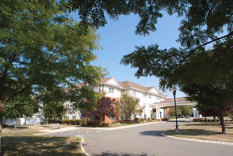 Exterior view of a multi-story senior living facility building with a covered entrance driveway, surrounded by trees and a well-maintained lawn under a clear blue sky.