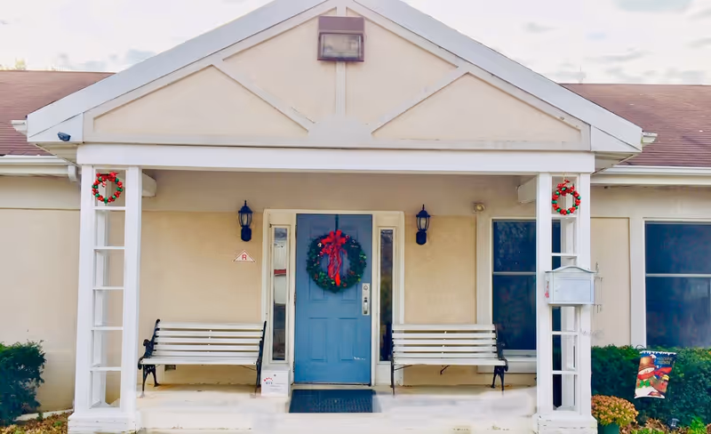 Covered building entrance with a blue door decorated with a wreath, two benches, and holiday wreaths on the porch columns.