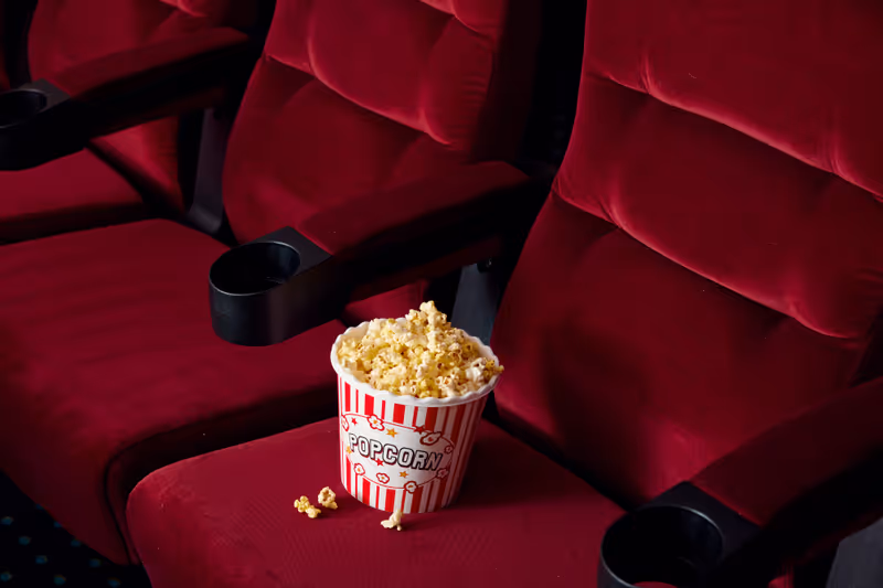 A bucket of popcorn placed on a red velvet theater seat with black armrests and cup holders, with a few pieces of popcorn spilled on the seat.