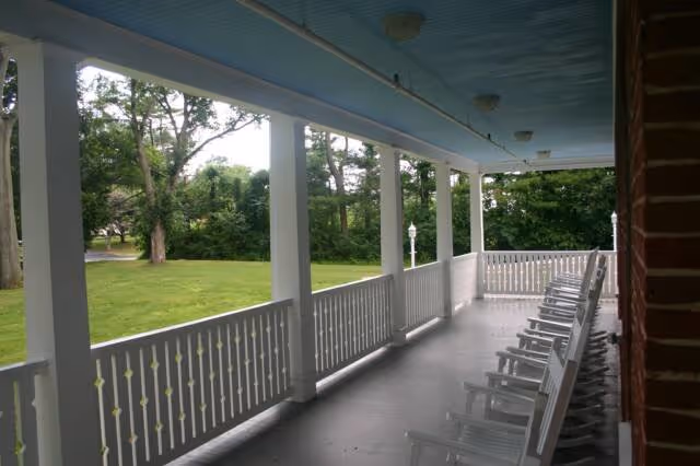 A covered porch with a row of white rocking chairs facing outward, overlooking a green lawn and trees. The porch has white railings and columns, with a light blue ceiling and a brick wall on one side.