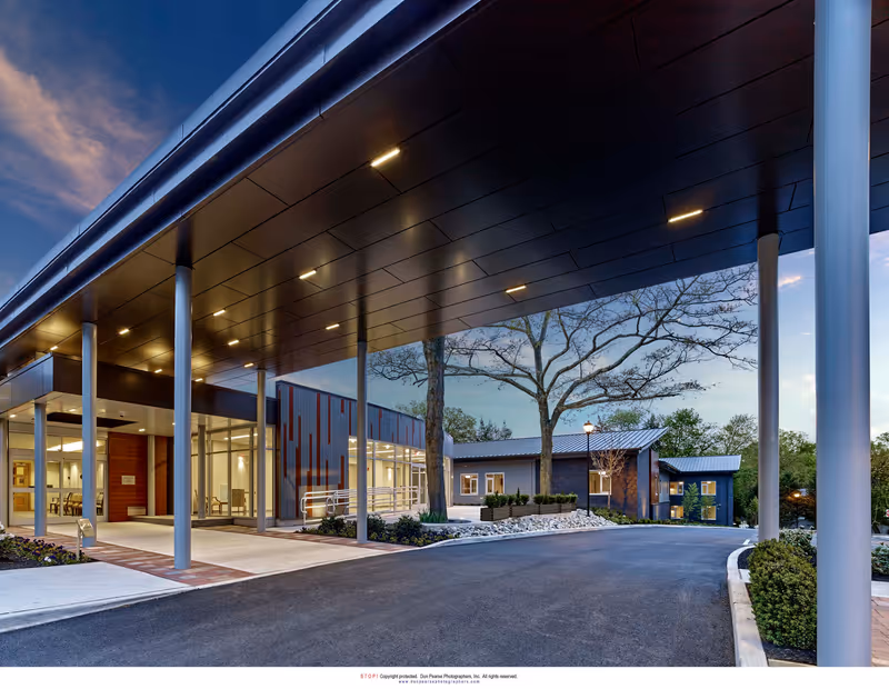 Exterior view of The Actors Fund Home building entrance at dusk, featuring a covered drop-off area with tall pillars, modern architecture, landscaped surroundings, and a clear sky with some clouds.