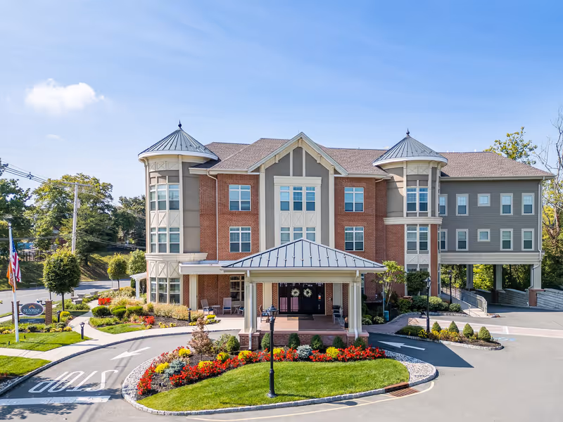 Front exterior of a three-story senior living building with a covered entrance, turret-style towers, and a landscaped circular driveway.