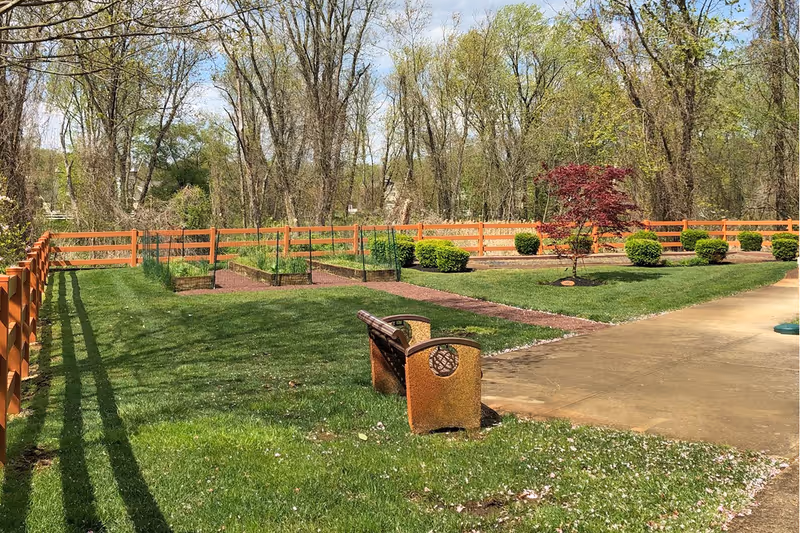 A peaceful outdoor garden area with a wooden bench on a grassy lawn, a paved walkway, raised garden beds with plants, neatly trimmed bushes, a small red-leafed tree, and a wooden fence surrounding the area with tall trees in the background under a partly cloudy sky.