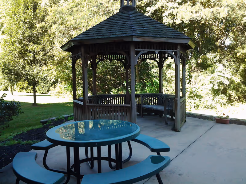 Outdoor patio area with a wooden gazebo featuring built-in benches and a round glass-top picnic table with attached benches on a concrete surface, surrounded by green trees and grass.
