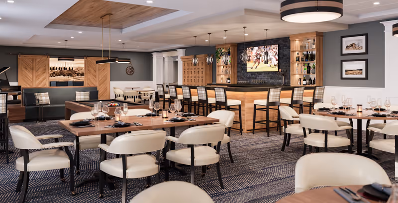 A modern dining room in a senior living facility featuring several round and rectangular wooden tables set with wine glasses, plates, and cutlery. Cream-colored cushioned chairs surround the tables. In the background, there is a bar area with high stools, shelves with bottles, and a large TV mounted on a dark stone wall. The room has a patterned carpet, soft lighting, and framed pictures on the walls.