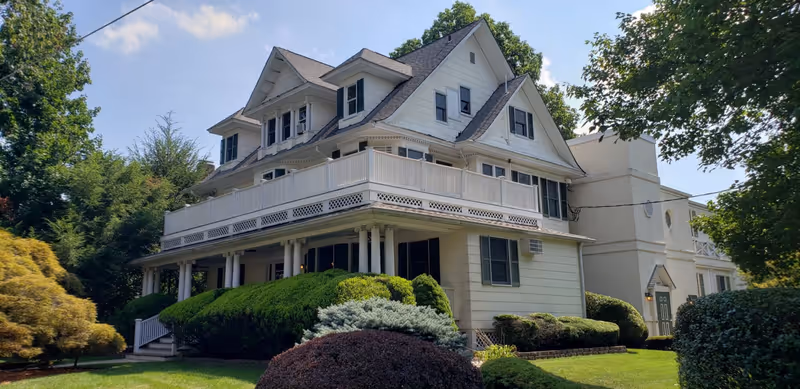 A large, multi-story white residential building with a wraparound porch and balcony, surrounded by well-maintained bushes and trees under a partly cloudy sky.