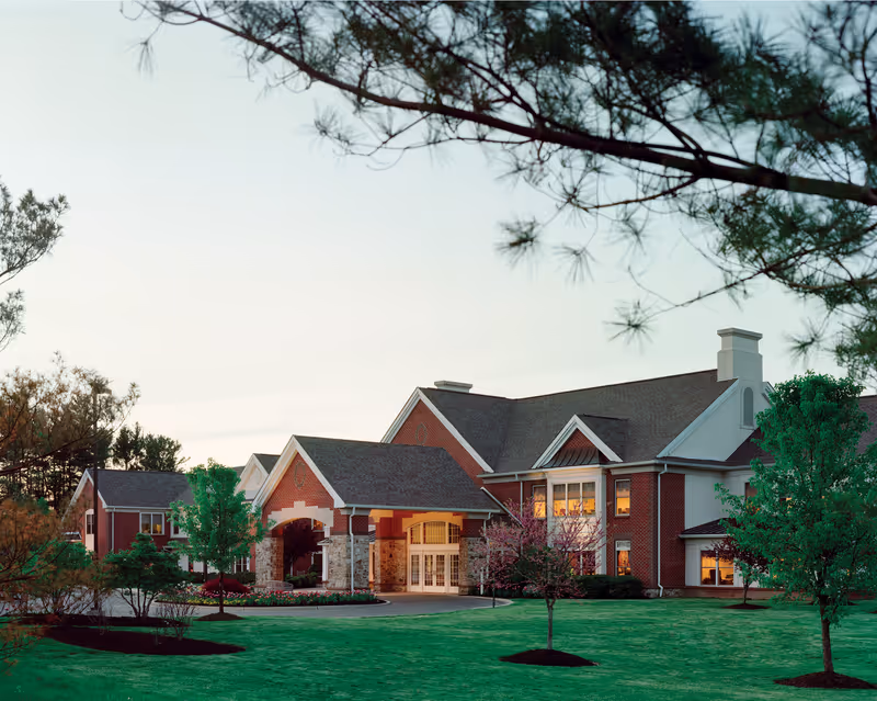 Exterior view of a large brick building with a covered entrance, surrounded by green grass, trees, and landscaping, under a clear sky at dusk.