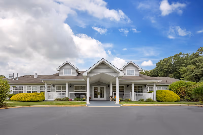 Front exterior view of a single-story building with a covered entrance supported by white columns, surrounded by green bushes and trees under a partly cloudy blue sky.