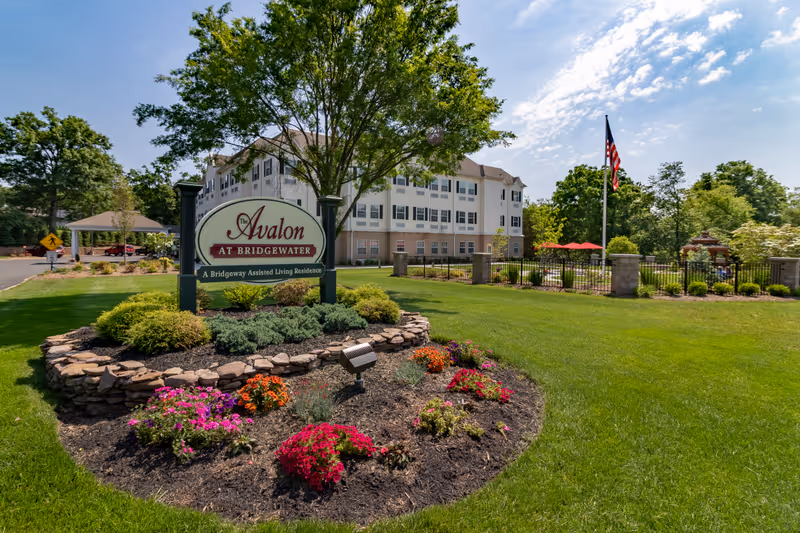 The exterior of Avalon Assisted Living at Bridgewater, showing a well-maintained lawn with colorful flower beds and shrubs. A large sign with the facility's name is prominently displayed in the foreground. In the background, there is a multi-story building with many windows, a flagpole with an American flag, and a gazebo surrounded by trees under a partly cloudy sky.