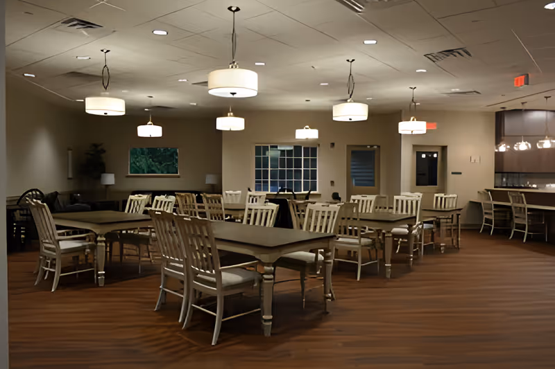 A spacious dining room with multiple wooden tables and chairs arranged neatly. The room has warm lighting from ceiling pendant lamps and a wooden floor. There is a counter with bar stools on the right side and windows on the far wall.