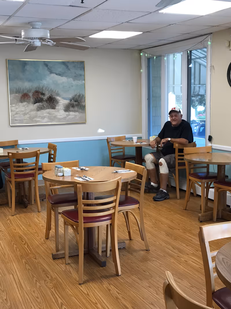 A dining room with wooden tables and chairs, a ceiling fan, a large painting of a beach scene on the wall, and a man wearing a black shirt, gray shorts, knee braces, and a white cap sitting at a table near a window.