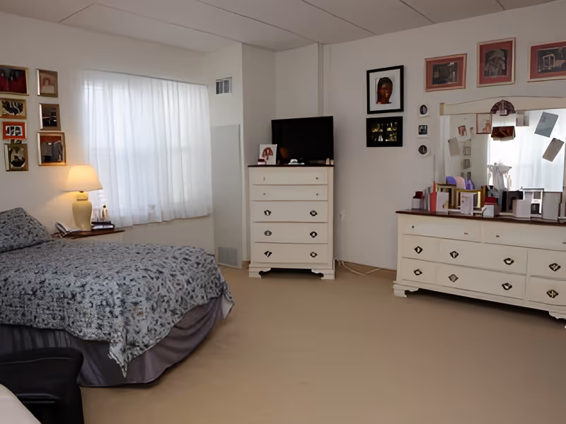 A bedroom with a bed covered in a patterned gray bedspread, a nightstand with a lamp and some items, a window with sheer white curtains, a white dresser with a TV on top, and a large white dresser with a mirror above it. Several framed pictures hang on the walls.