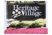 Entrance sign that reads 'Heritage Village' surrounded by shrubs and bright pink flowers.
