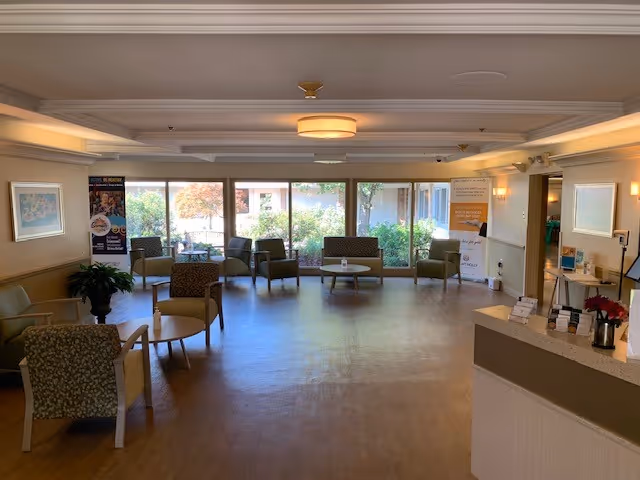 A spacious and well-lit common area in a senior living facility with multiple chairs and small tables arranged around the room. Large windows at the back provide a view of greenery outside. The room has light-colored walls and ceiling with modern light fixtures, and there is a reception or information desk on the right side.