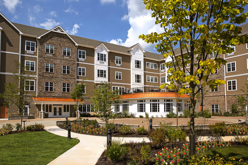Exterior view of a multi-story senior living facility building with a combination of stone and siding facade. The building is surrounded by landscaped gardens with trees, shrubs, and flower beds. A paved walkway curves through the garden area under a partly cloudy blue sky.