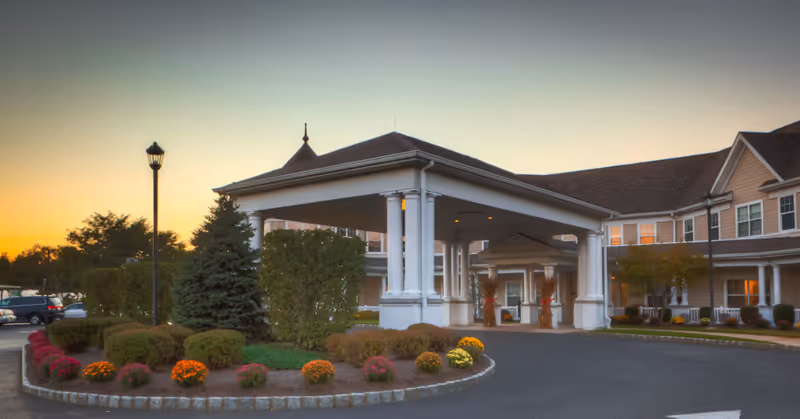 Front entrance of a senior living facility with a covered porte-cochere, landscaped circular planter, and a warm evening sky.