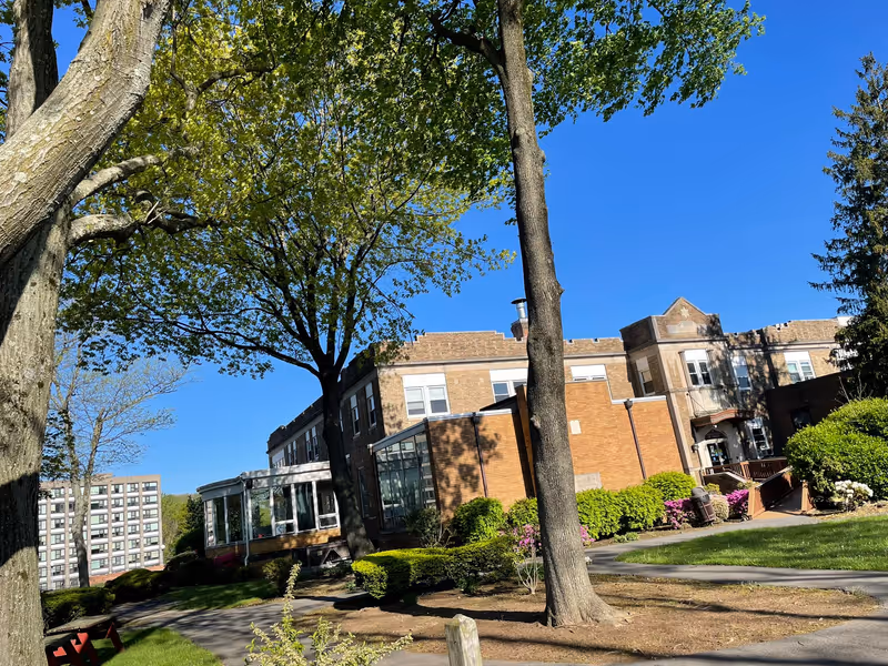 Exterior view of a multi-story brick building with large windows, surrounded by green trees, bushes, and a well-maintained lawn under a clear blue sky.