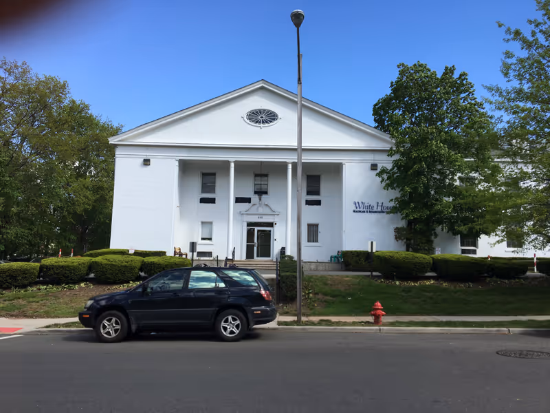 Front exterior view of a white two-story building with classical columns and a triangular pediment. There is a black car parked on the street in front of the building, a streetlamp in the center, and green bushes and trees surrounding the building. The sign on the building reads 'White House Healthcare Center'.