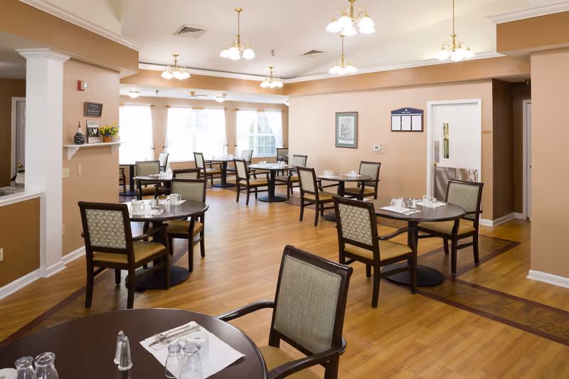 A bright dining room in Maurice House Assisted Living Community with several round tables and chairs arranged neatly. Each table is set with glasses, napkins, and silverware. The room has wooden flooring, beige walls, multiple ceiling light fixtures, and large windows with white curtains allowing natural light to fill the space.