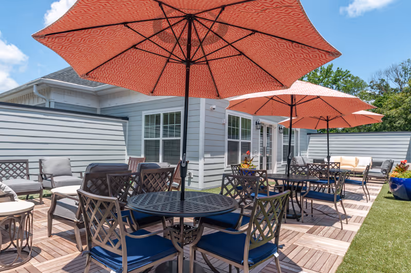 Outdoor patio area with multiple round metal tables and chairs with blue cushions, shaded by large red patterned umbrellas. The patio has wooden tile flooring and is adjacent to a light gray building with white-trimmed windows and doors. There are also cushioned lounge chairs and large blue planters with flowers along the edge.