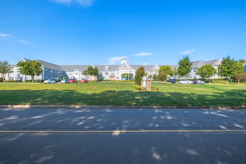Wide exterior view of Sunnyside Manor, a large two-story senior living facility with a gray roof and light-colored siding. The building is surrounded by green lawns, trees, and a parking area with several cars. The sky is clear and blue.